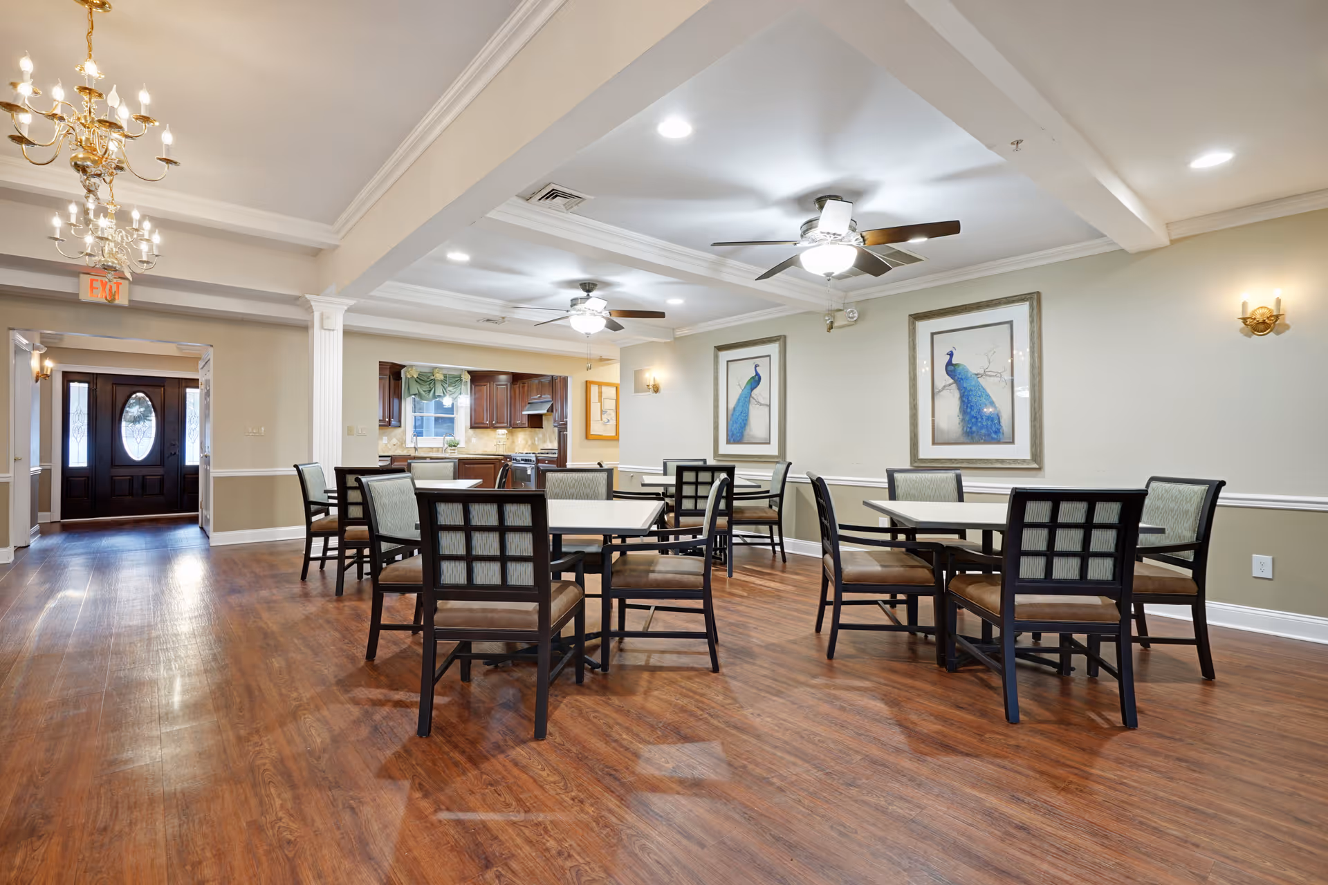 A well-lit dining area in a senior living facility with several square tables and chairs arranged on a wooden floor. The room features ceiling fans with lights, framed peacock artwork on the walls, and a chandelier near the entrance. In the background, there is a kitchen area with dark wood cabinets and a window with a valance.