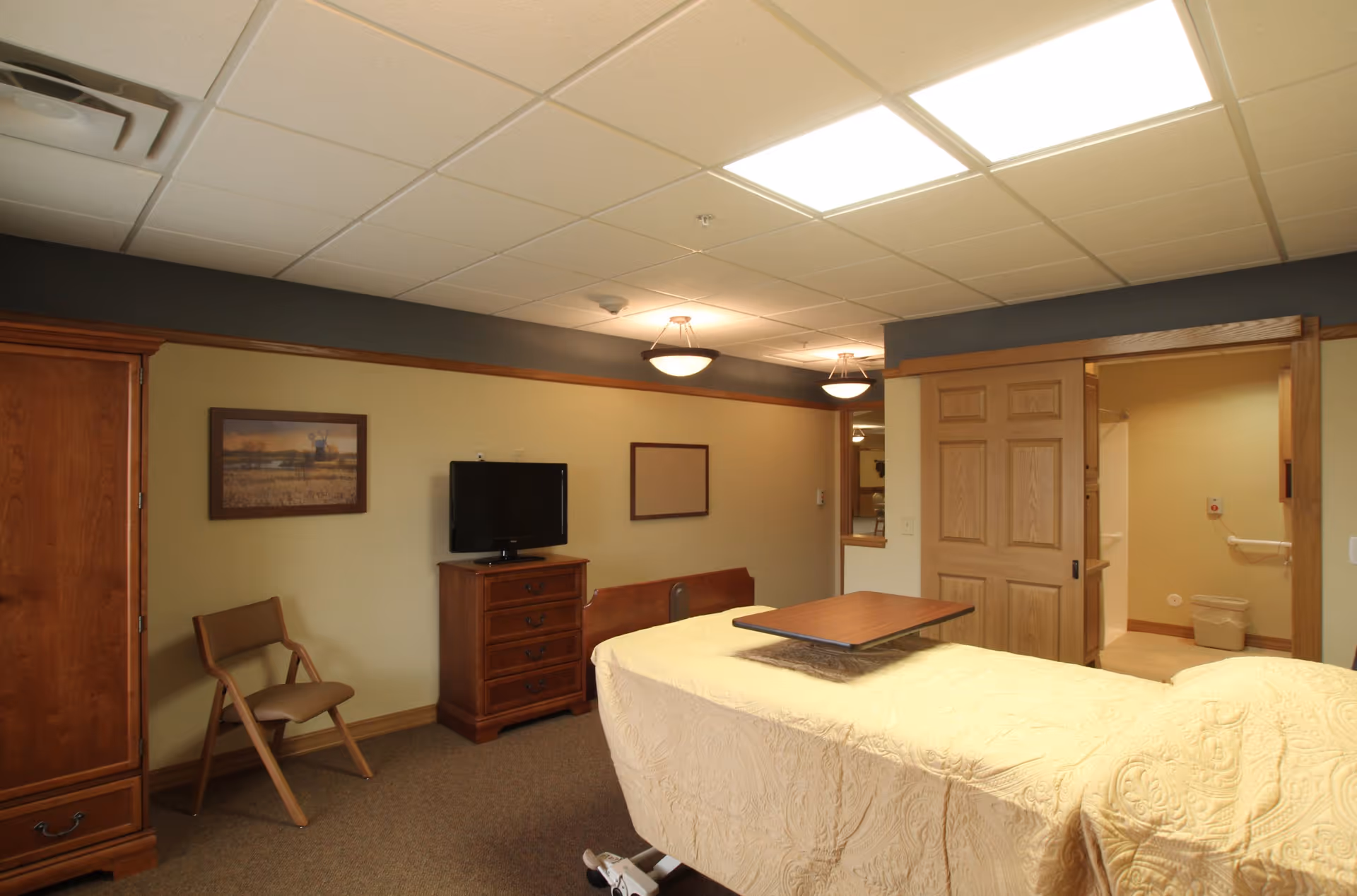 A senior living facility bedroom with a hospital-style bed covered in a cream quilt, a wooden overbed table, a wooden dresser with a flat-screen TV on top, a wooden chair, and a framed picture on the wall. The room has beige walls with a dark gray accent near the ceiling and two ceiling lights. A sliding wooden door leads to an adjacent bathroom area.