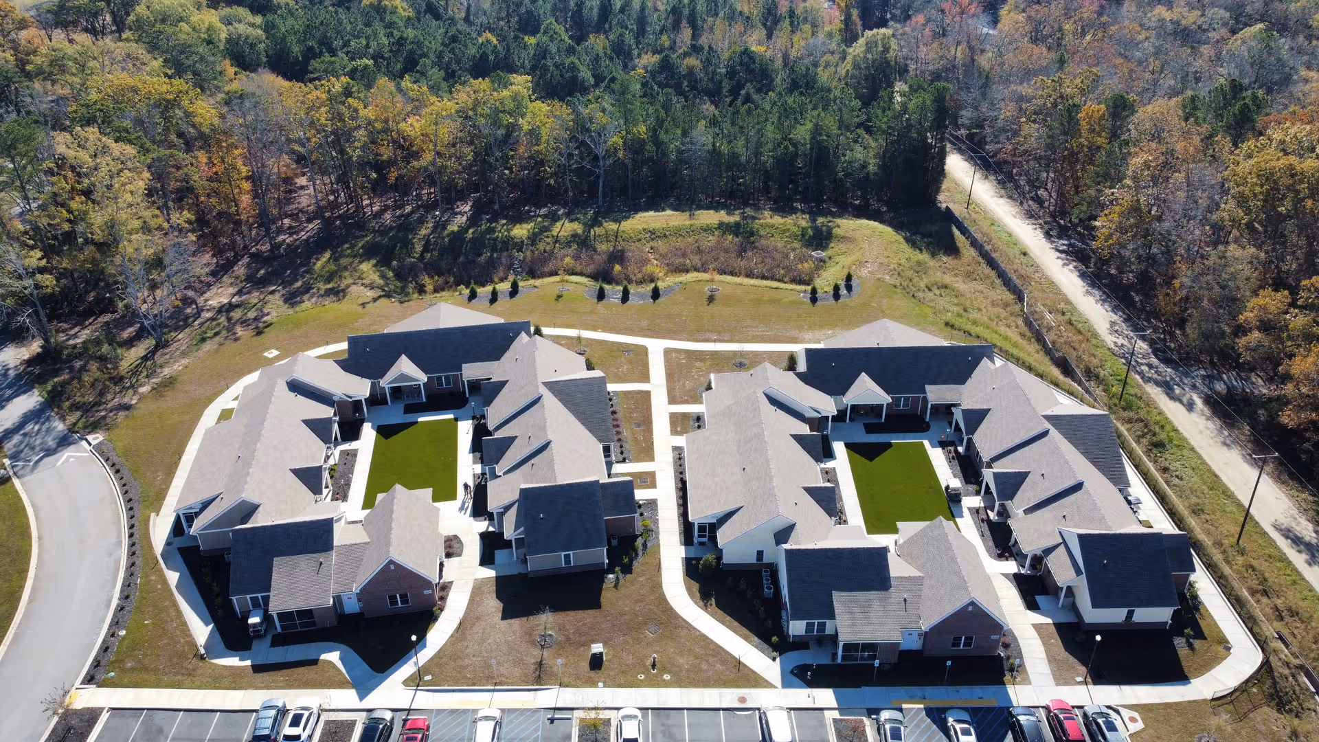 Aerial view of The Heritage At Lowman senior living facility showing two large, U-shaped buildings with gray roofs and green courtyards in the center. The buildings are surrounded by paved walkways, parking spaces with several cars, and bordered by trees and greenery.