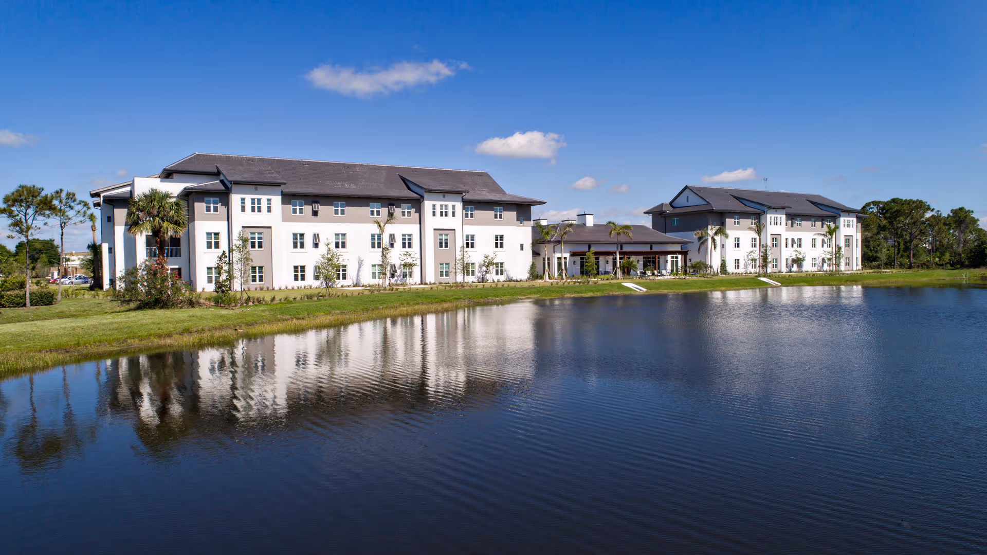 A three-story light-colored senior living building along a lakeshore with its reflection in the water under a clear blue sky.
