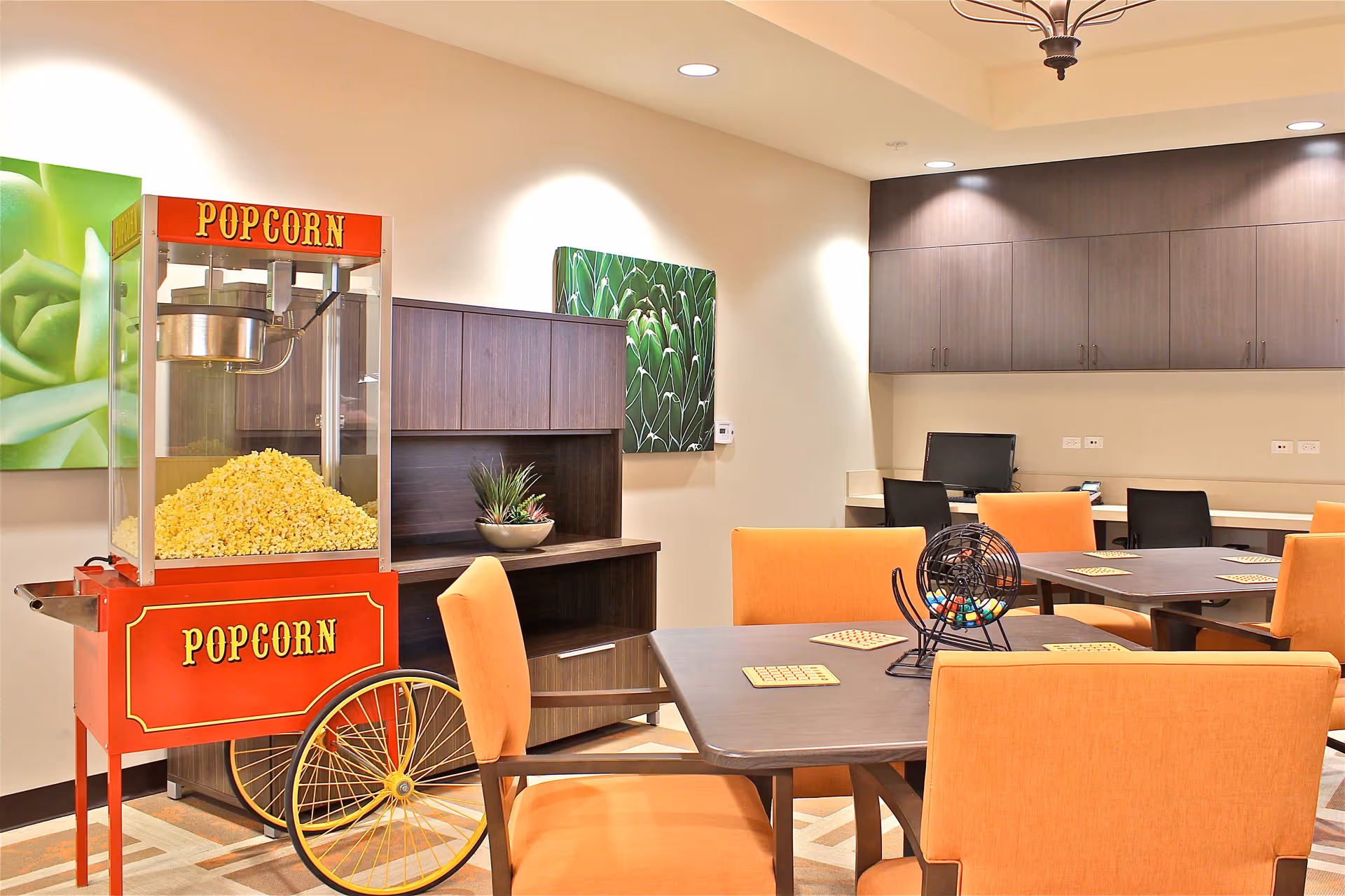 A bright room with orange chairs around dark wooden tables, each table set with bingo cards and a bingo cage. A red popcorn machine filled with popcorn is positioned near the wall, which is decorated with green plant artwork. There are cabinets and a computer workstation in the background.