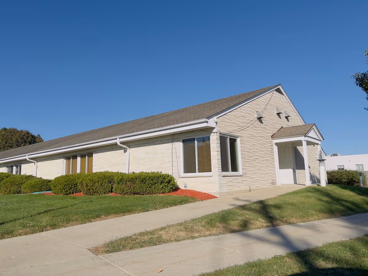 Single-story beige brick building with a small portico entrance, manicured shrubs, lawn and a sidewalk under a clear blue sky.