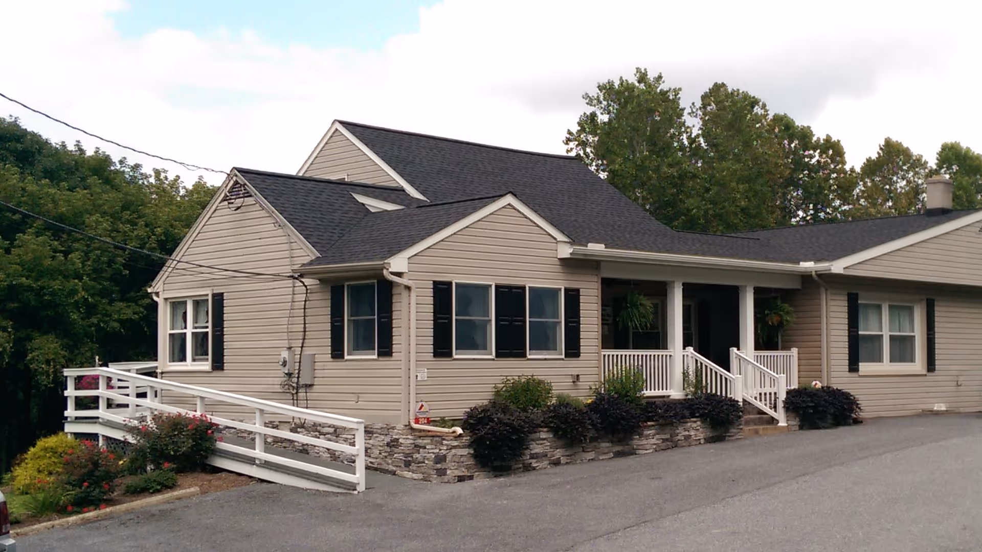 Exterior view of a single-story beige building with black shutters and a dark shingled roof. The building has a white railing along a ramp and steps leading to a covered porch entrance. There are bushes and plants along the front of the building and trees in the background under a partly cloudy sky.