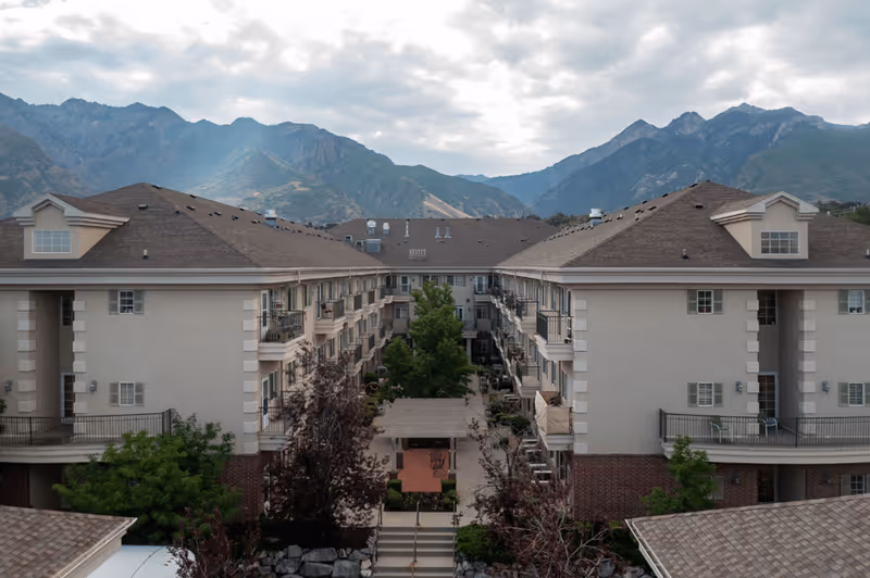 Exterior view of a multi-story senior living facility with balconies, surrounded by trees and landscaping, set against a backdrop of mountains under a cloudy sky.