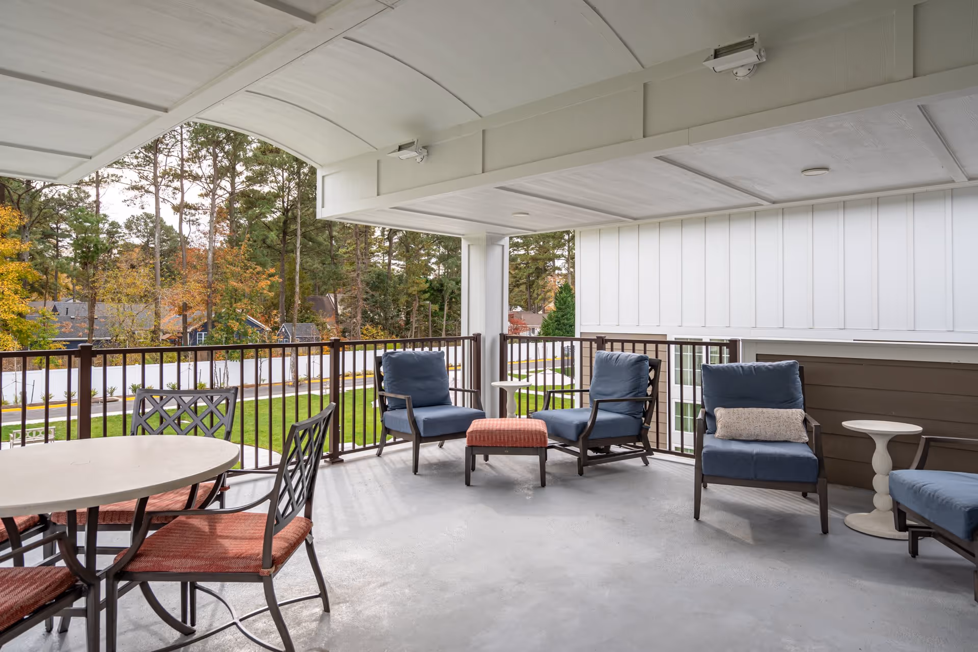 Covered outdoor patio/balcony with cushioned chairs, a table and ottoman overlooking a landscaped yard and trees.