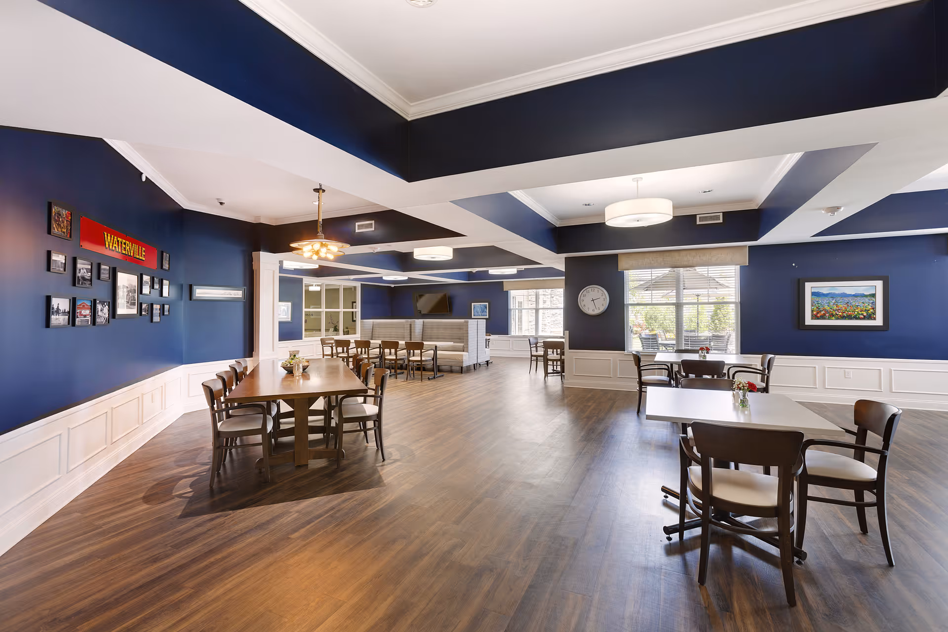A spacious dining room with dark blue walls and white wainscoting, featuring multiple wooden tables and chairs arranged throughout the room. The room has large windows allowing natural light to enter, a wall clock, framed pictures, and a sign that reads 'WATERVILLE'. The floor is wooden, and the ceiling has recessed lighting and modern light fixtures.