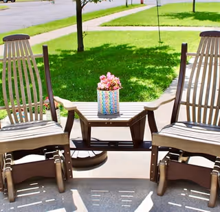 Two wooden outdoor chairs with armrests and a small table between them holding a colorful pot with pink flowers, set on a concrete patio with green grass and trees in the background.