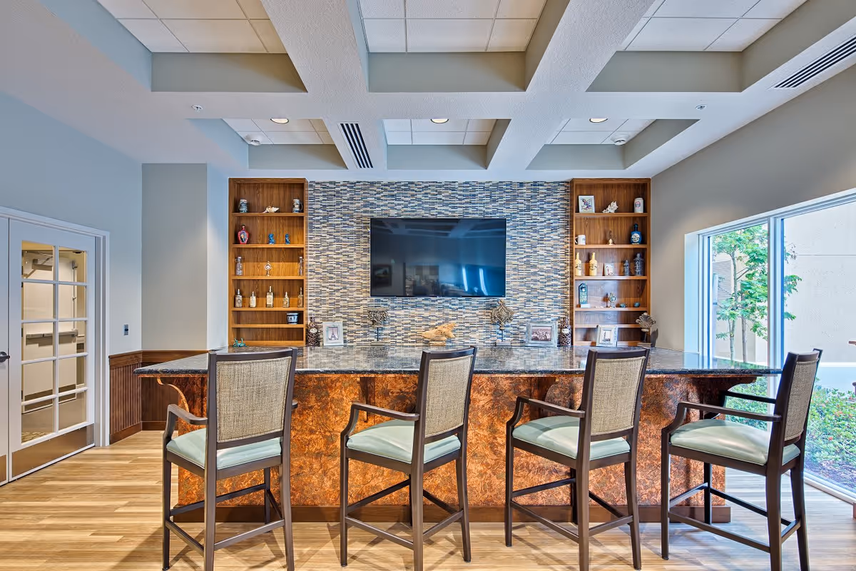 Interior view of a senior living facility lounge area with a large flat-screen TV mounted on a tiled wall. Below the TV is a long counter with a marble-like surface and four high chairs with armrests and cushioned seats. On either side of the TV are wooden shelves displaying decorative items and framed photos. To the right, large windows provide a view of greenery outside. The ceiling has recessed lighting and a coffered design.