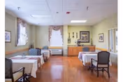 Small dining room with several tables draped in white tablecloths, upholstered chairs, windows with curtains, and a sideboard along the far wall.