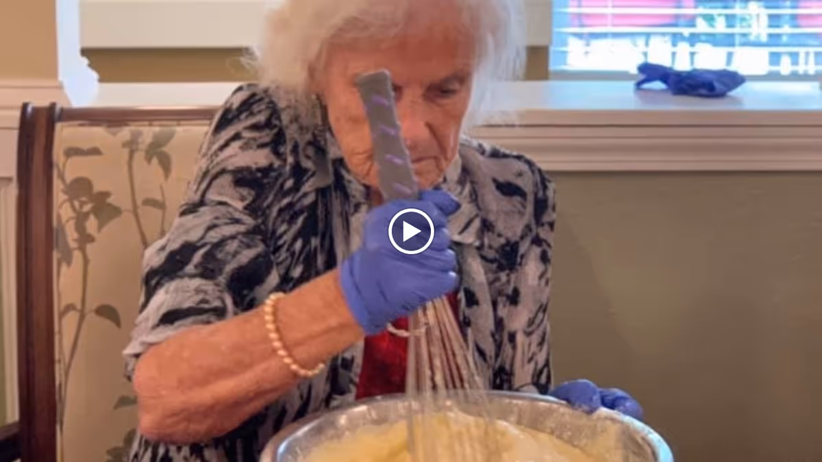 An elderly woman wearing a patterned shirt and blue gloves is sitting at a table, whisking batter in a large metal bowl. She is focused on the task, and there is a window with blinds in the background.