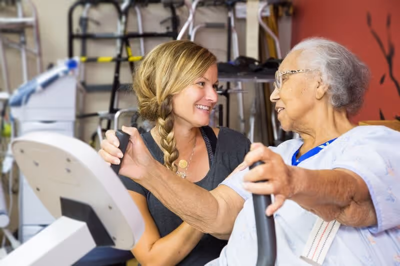An elderly woman using a seated exercise machine while a younger woman with a braid smiles and supports her, in a room with exercise equipment and walkers in the background.