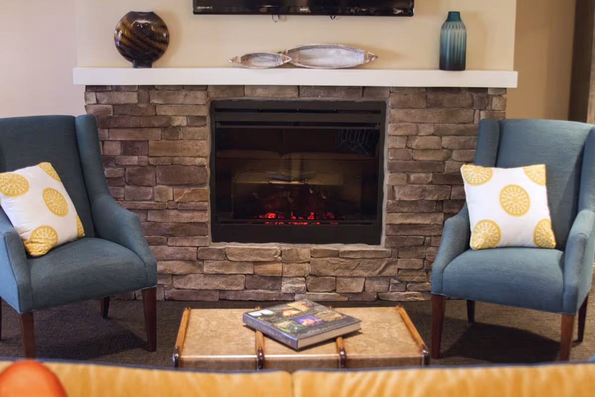 A cozy living room area with a stone fireplace in the center, flanked by two blue armchairs each with a white pillow featuring yellow circular patterns. A small table with a book on it is placed in front of the fireplace. Decorative items are on the mantel above the fireplace.
