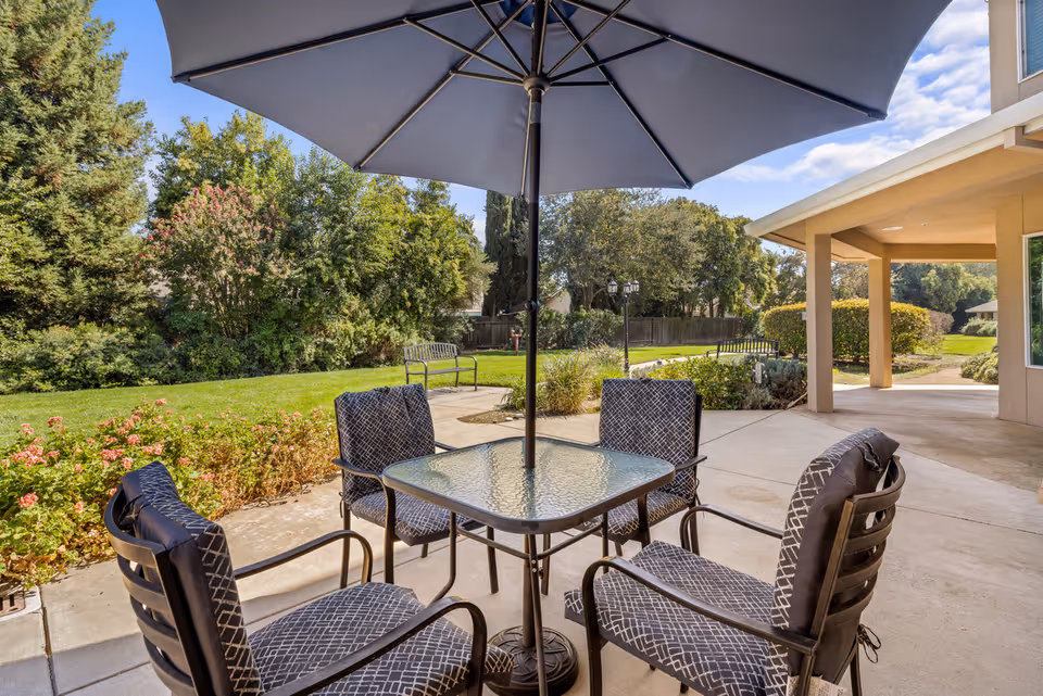 Outdoor patio with a glass table, umbrella and four cushioned chairs overlooking a grassy garden beside the building.