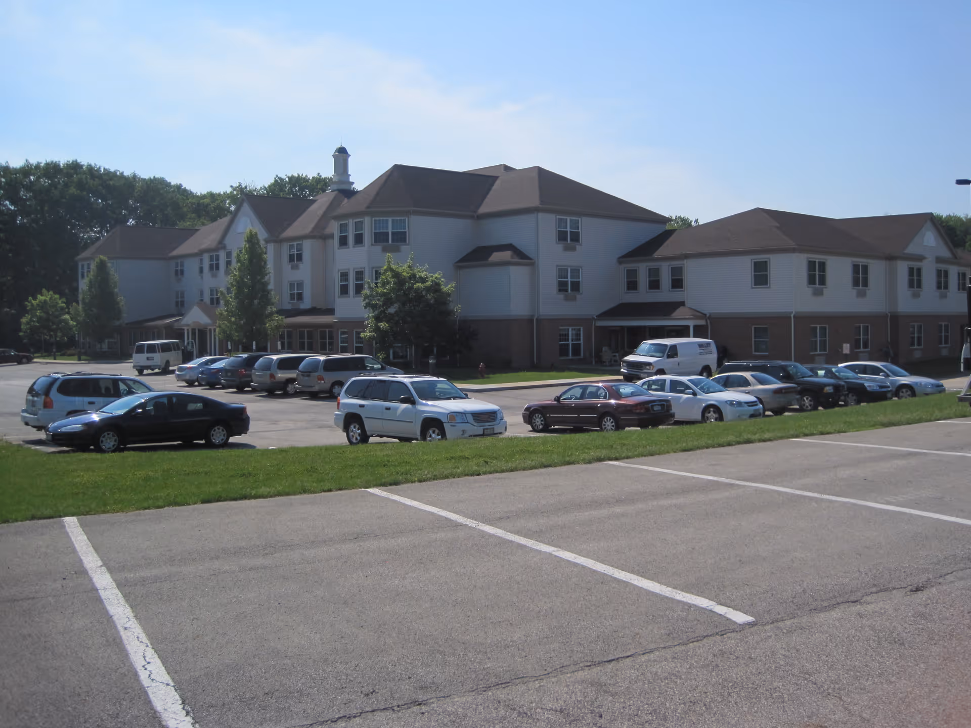 Exterior view of a large, multi-story assisted living facility building with a parking lot in front containing several parked cars. The building has a combination of brick and light-colored siding with multiple windows and a pitched roof. Trees and greenery surround the area under a clear blue sky.