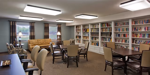A well-lit room with multiple tables and chairs arranged for group seating, surrounded by bookshelves filled with books. There are two windows with curtains, two table lamps, and additional seating including a sofa and armchairs. The room has a cozy and inviting atmosphere.