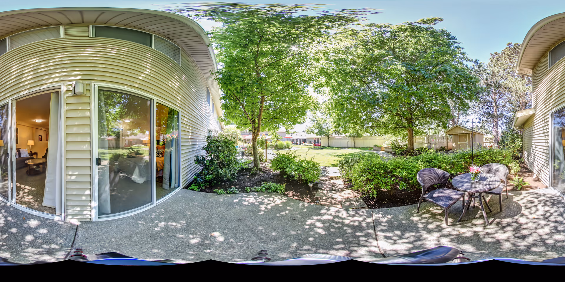 Outdoor patio area at Aegis Living Lynnwood with a small round table and two chairs on a concrete surface. The patio is surrounded by green bushes and trees providing shade. Sliding glass doors lead inside the building where a living room is partially visible. The area opens up to a grassy lawn with more trees and a small shed in the background under a clear blue sky.