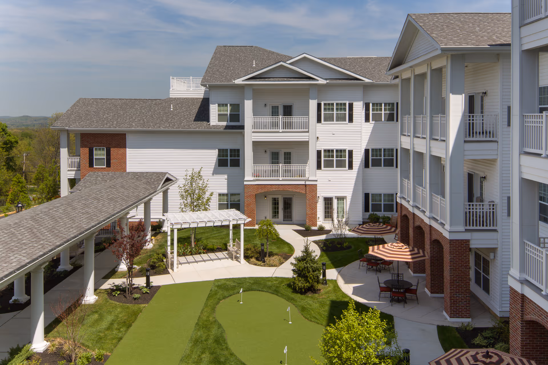 Outdoor courtyard area of a senior living facility with a putting green, landscaped garden, white pergola, and patio tables with striped umbrellas. The building surrounding the courtyard is three stories tall with white siding, brick accents, balconies, and a covered walkway.