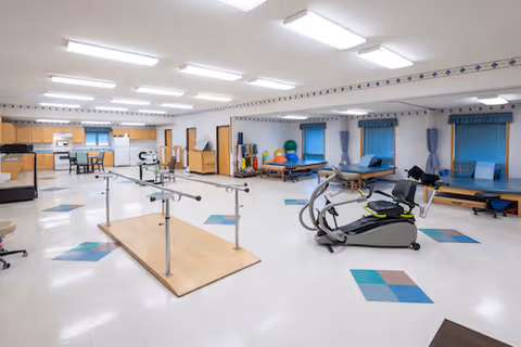 A spacious rehabilitation room with exercise equipment including parallel bars, an exercise bike, therapy tables, and various therapy balls. The room has bright overhead lighting, windows with blue blinds and curtains, and a kitchenette area with cabinets and appliances in the background.