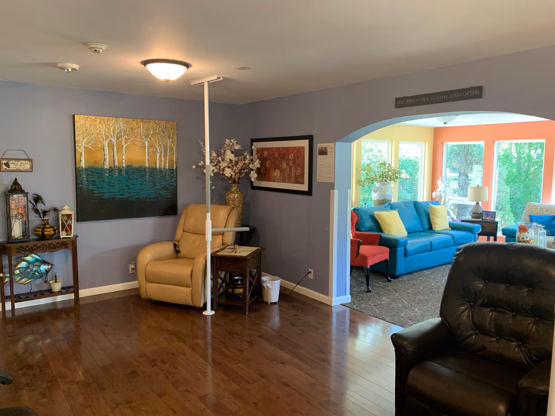 Interior view of a senior living facility common area with wooden flooring, a beige recliner chair with a support pole, decorative paintings on the walls, and a small table with flowers. Through an archway, a bright sitting area is visible with a blue couch, red chairs, yellow pillows, large windows, and a lamp on a side table.