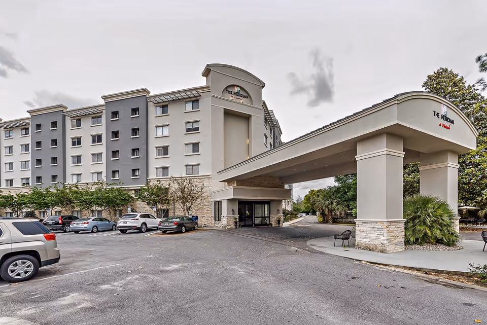Exterior view of a multi-story building with a covered entrance and parking lot in front. Several cars are parked, and there are trees and shrubs around the building. The building has a sign that reads 'The Berkman at Westwood.'