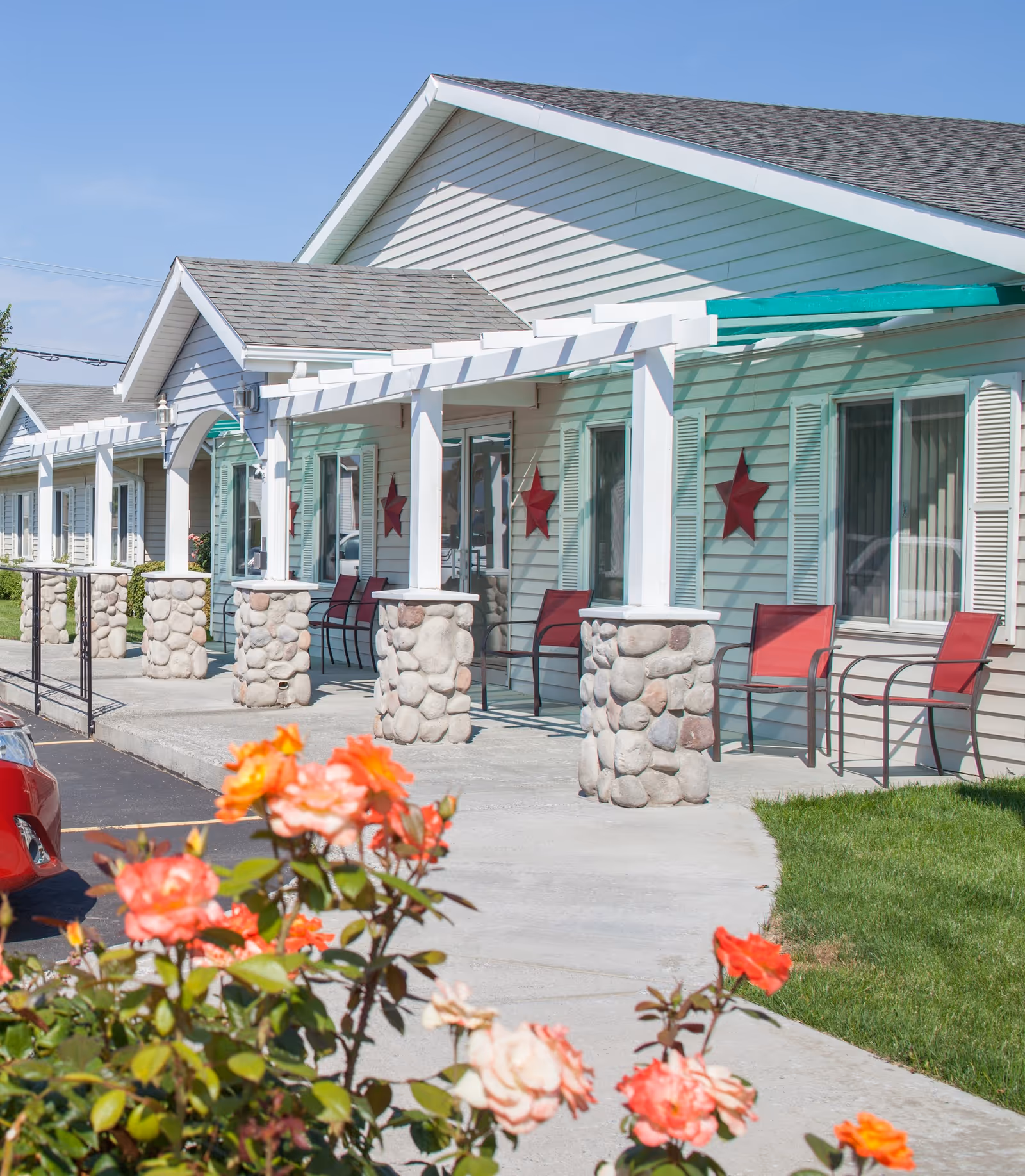 Exterior view of Monroe House, A Senior Living Community, showing a single-story building with light-colored siding, stone pillars supporting a white pergola, red star decorations on the walls, red chairs arranged along the front porch, and blooming orange and pink flowers in the foreground.