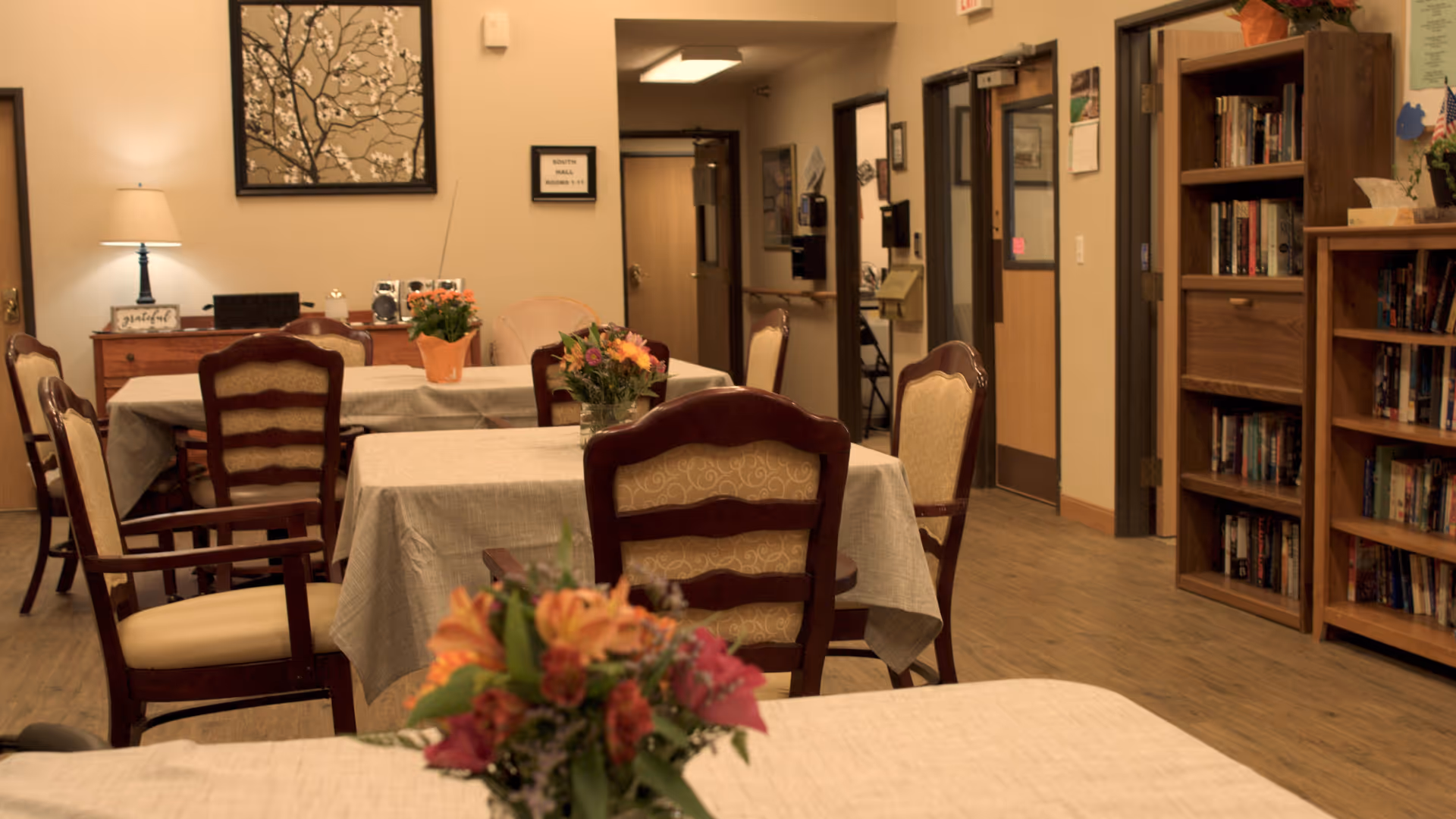 A cozy common area with several tables covered in beige tablecloths, each adorned with a vase of colorful flowers. Wooden chairs with cushioned seats surround the tables. In the background, there is a wooden bookshelf filled with books, a small lamp on a side table, and a framed floral artwork on the wall. The room has warm lighting and a wooden floor, with a hallway visible in the back.