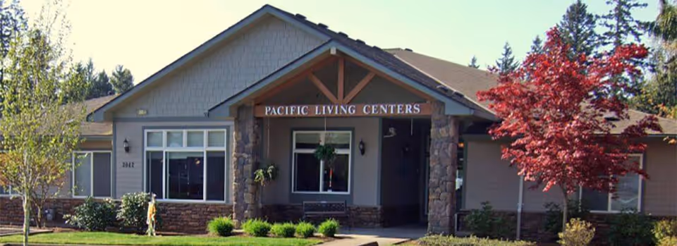 Front exterior view of a single-story building with a peaked roof and stone pillars at the entrance. The building has a sign that reads 'PACIFIC LIVING CENTERS' above the entrance. There are trees and shrubs in the landscaped area in front of the building.