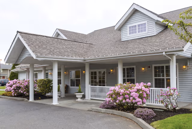 Exterior view of Avista Senior Living Ferndale showing a single-story building with a covered entrance supported by white columns. There are blooming pink and purple flowers and shrubs along the front, with a paved driveway and green lawn visible.
