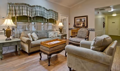 A cozy senior living facility common area with a beige sofa, two armchairs, a wooden coffee table, and two side tables with lamps. The room has wooden flooring, large windows with green and beige valances, and a framed picture on the wall.