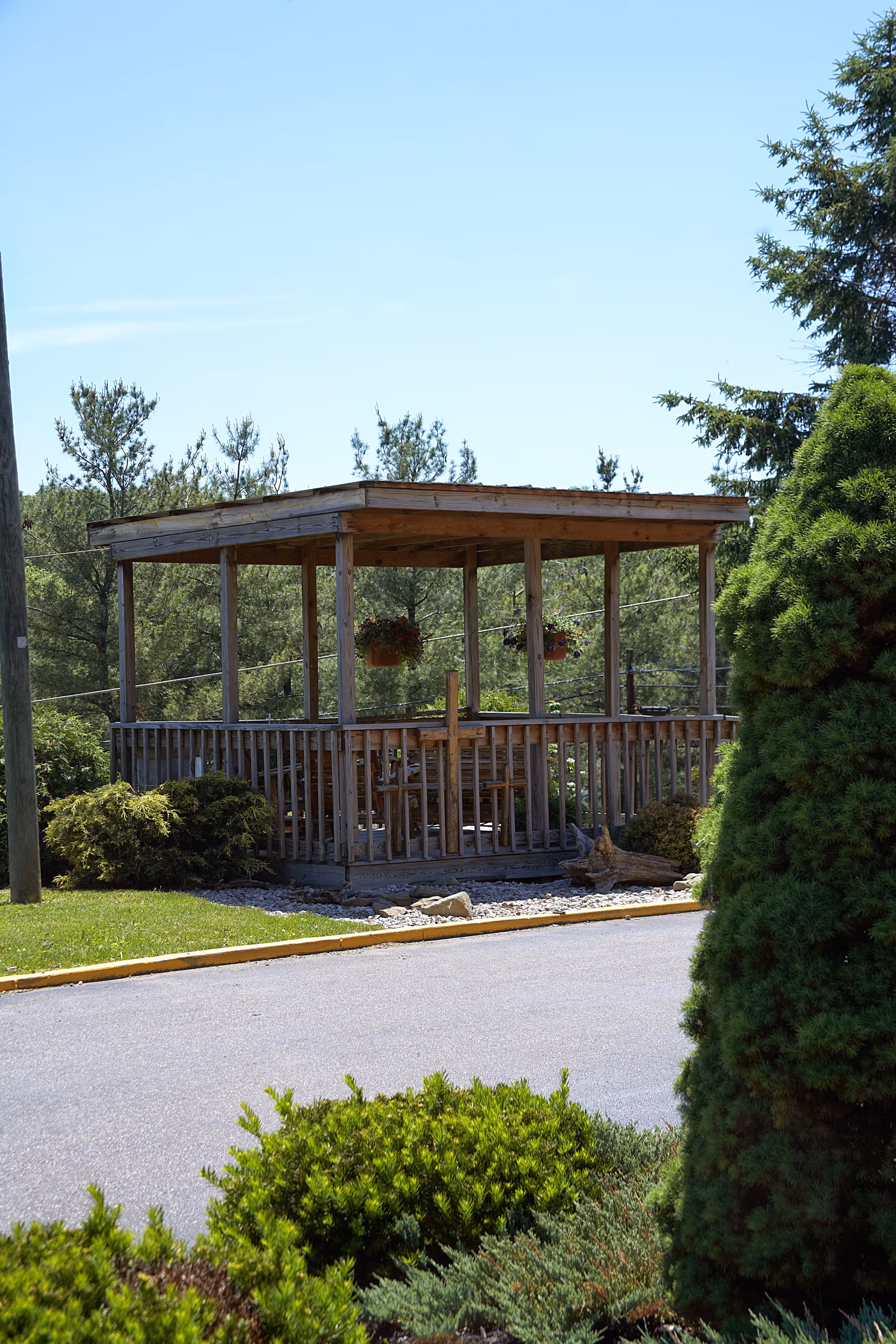 A wooden gazebo with a roof and open sides surrounded by greenery, including bushes and trees, situated near a paved road under a clear blue sky.