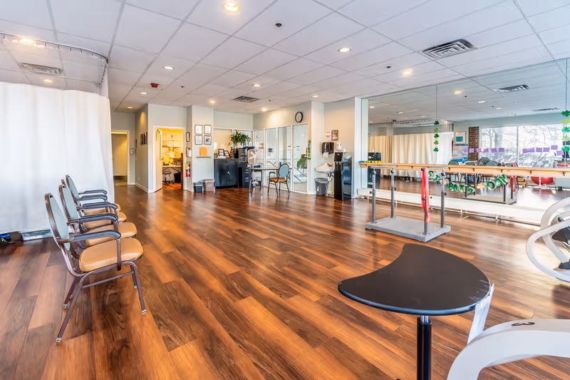 A spacious room with wooden flooring and a row of chairs along one wall. The room has a mirrored wall with a ballet barre and exercise equipment, suggesting it is used for physical therapy or exercise. There is a desk area with chairs and office supplies in the background, along with a white curtain divider and large windows letting in natural light.