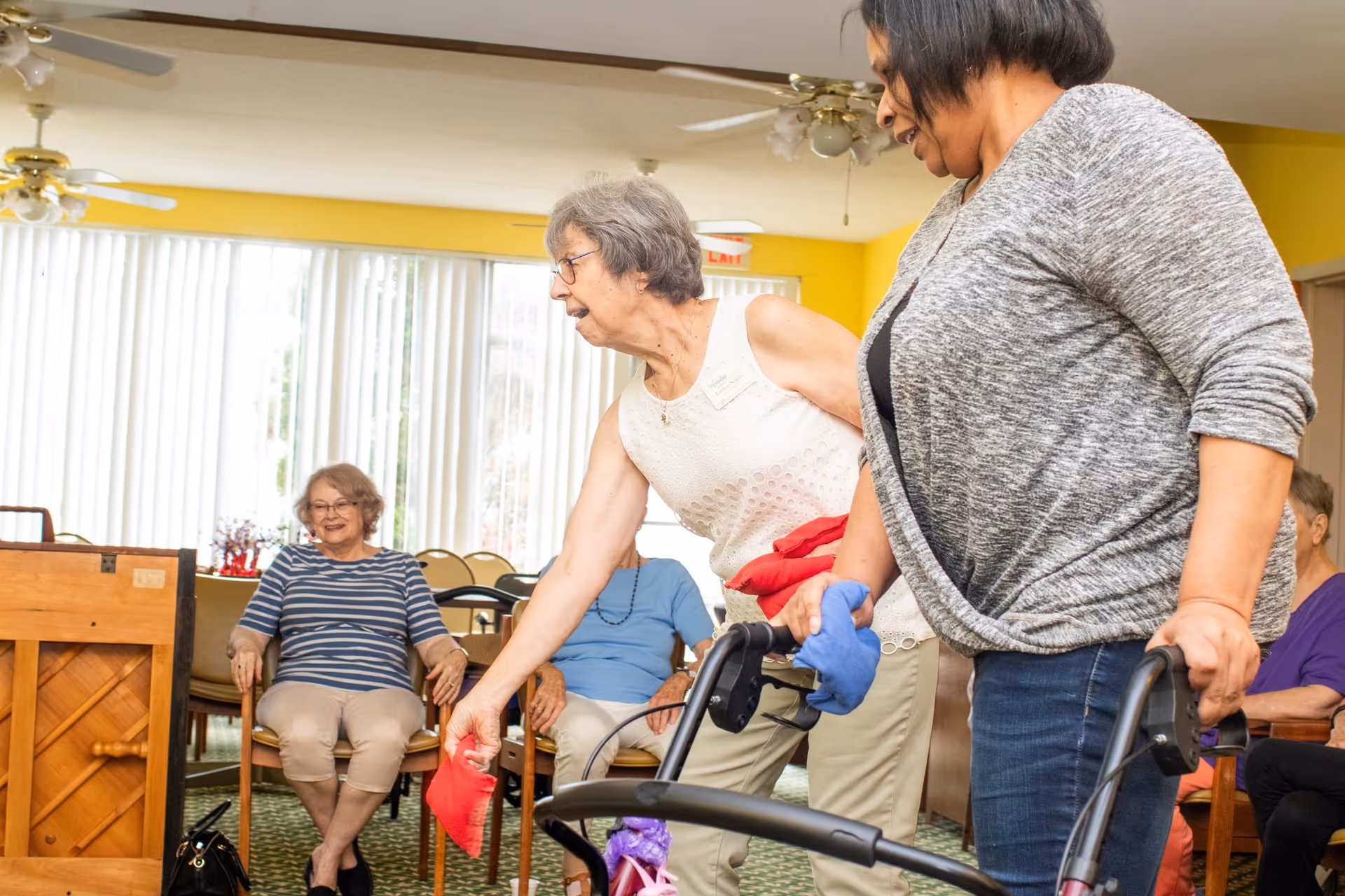 Two women standing and holding bean bags, one using a walker, participating in an activity in a senior living community room with other seated seniors in the background.
