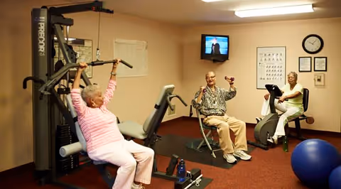 Three elderly individuals exercising in a fitness room. One woman is using a weight machine, a man is lifting small dumbbells while seated, and another woman is on a stationary bike. The room has beige walls, a wall-mounted TV, exercise posters, a clock, and a blue exercise ball on the floor.