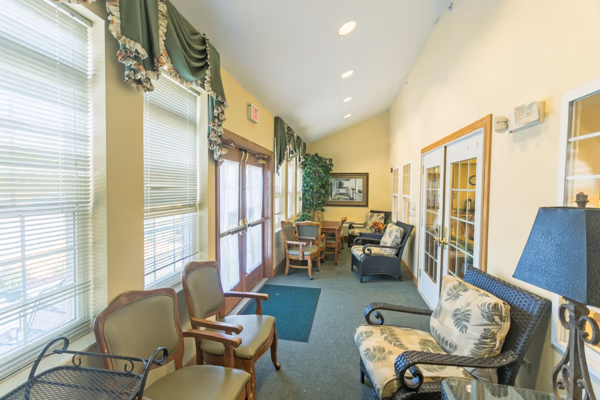 A bright, narrow sitting area with multiple chairs and small tables along the walls. Large windows with blinds and green valances let in natural light. There is a double door with glass panels and an exit sign above it. The walls are painted light yellow, and the floor is carpeted in green. A framed picture and a tall plant decorate the far end of the room.