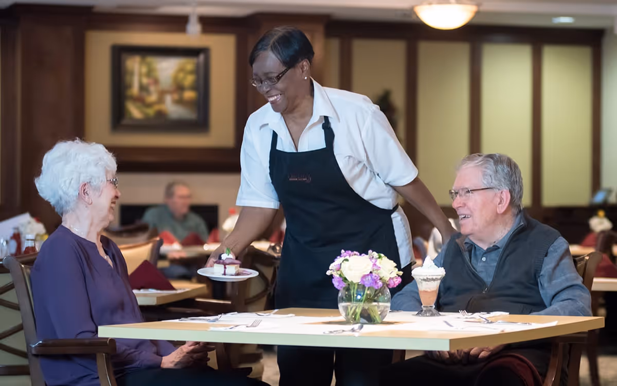 A smiling waitress serving a dessert plate to an elderly woman seated at a dining table, with an elderly man sitting next to her. The table has a small vase with flowers and a dessert glass. The setting is a warm, well-lit dining room with other diners in the background.