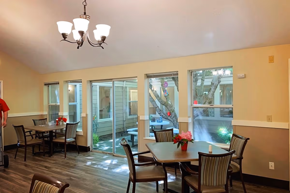 A dining area with wooden tables and chairs, a chandelier hanging from the ceiling, large windows and a sliding glass door looking out to a courtyard with a tree and outdoor seating. There are small flower arrangements on the tables.