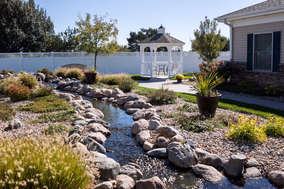 Landscaped outdoor courtyard with a rock-lined stream leading to a white gazebo beside a residential building.
