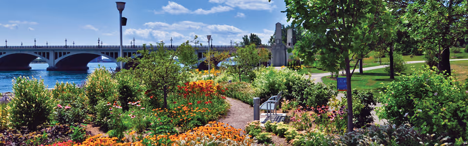 A colorful riverside garden with a winding path, flowers, a bench, and a distant bridge.