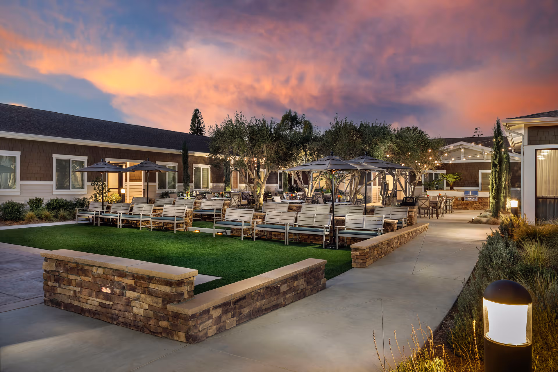 Outdoor courtyard at sunset with rows of benches, umbrellas, string lights, and walkways around a grassy area.