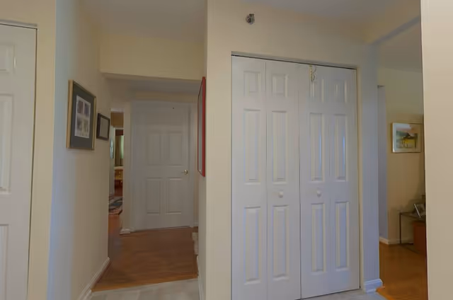 Interior view of a hallway in a residential facility with beige walls and white doors. There is a closet with double white doors in the foreground, a framed picture on the left wall, and a glimpse into other rooms with wooden flooring.