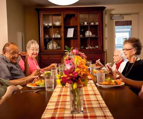Several elderly residents sit around a dining table sharing a meal with a floral centerpiece in a warmly lit dining room.