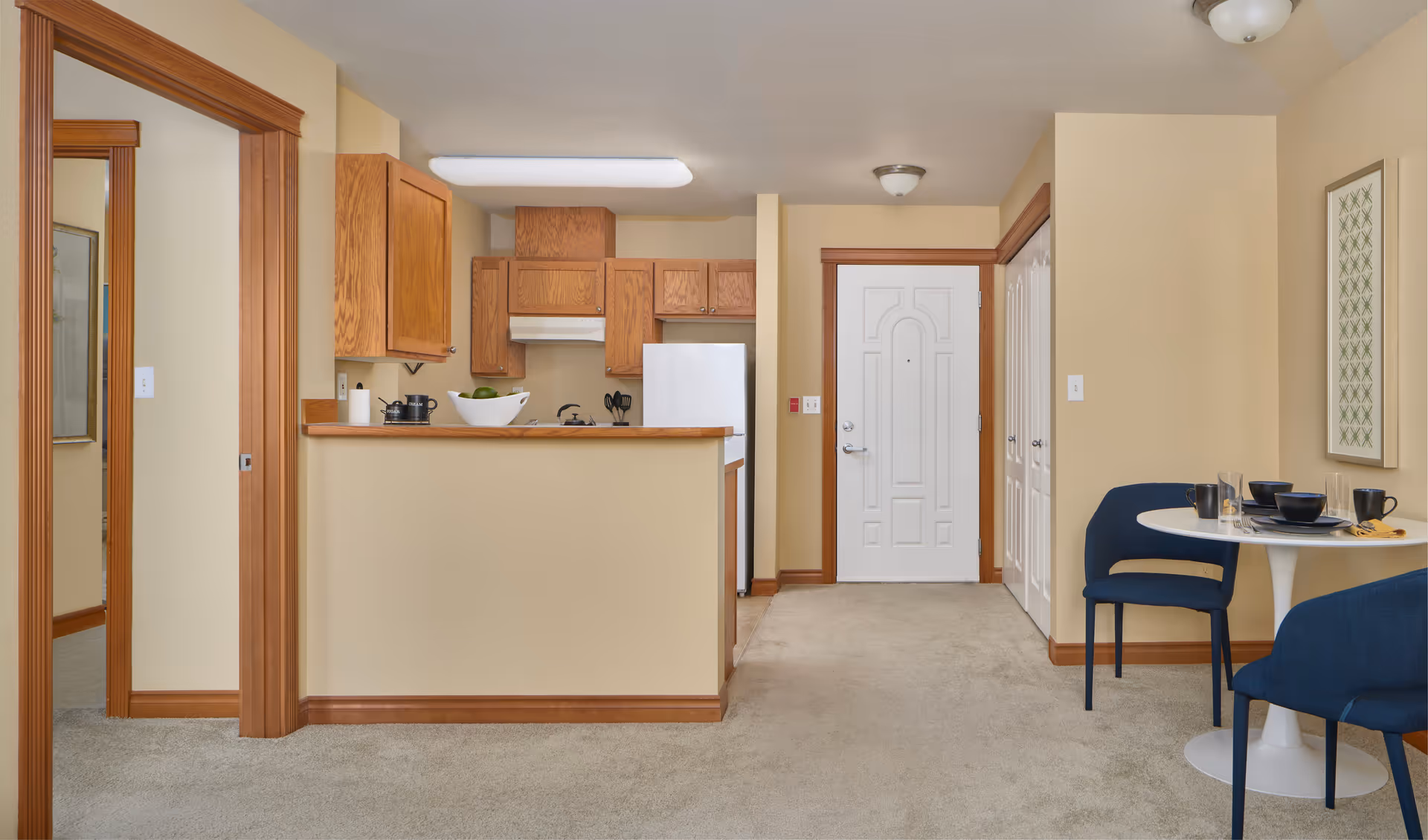 Interior view of a senior living apartment showing a small kitchen with wooden cabinets, a white refrigerator, and a counter with a bowl of green apples. To the right is a white front door with a small dining area featuring a round white table set with black dishes and blue chairs. The walls are painted beige and the floor is carpeted.