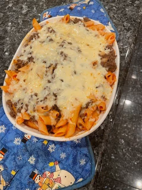 Baked penne pasta with meat and melted cheese in a white oval dish on a blue snowman-patterned pot holder atop a dark granite countertop.