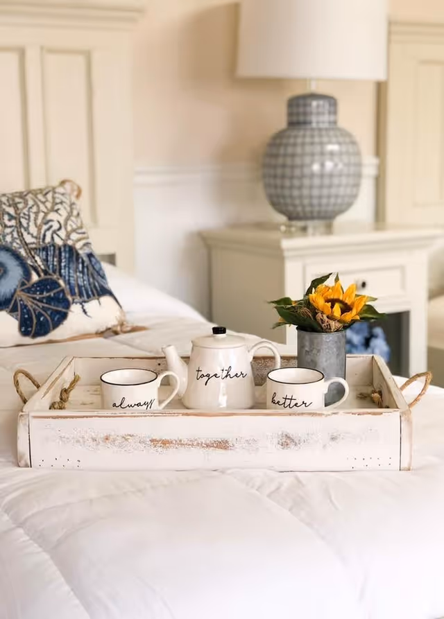 A white wooden tray with two white mugs and a teapot labeled 'always', 'together', and 'better' respectively, along with a small metal vase holding a sunflower, placed on a white bedspread in a cozy bedroom setting with a bedside table and lamp in the background.