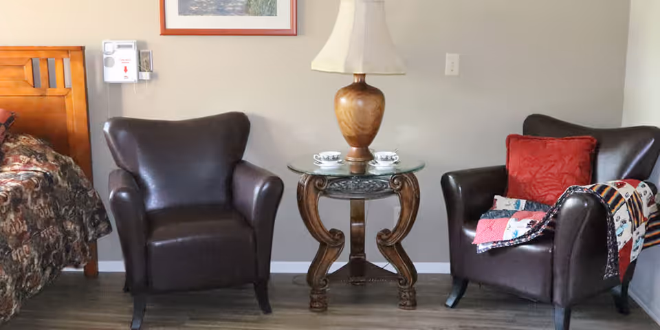 A cozy seating area in a room with two dark brown leather armchairs separated by a small round wooden table with a glass top. On the table is a wooden lamp with a beige lampshade and two teacups with saucers. To the left, part of a wooden bed frame with a floral bedspread is visible. The floor is wooden, and the walls are painted light gray.