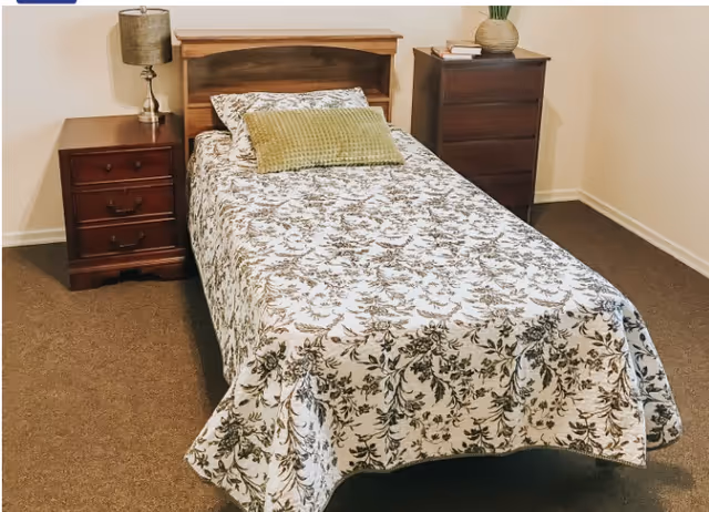 Twin bed with a floral bedspread flanked by a nightstand with lamp and a tall dresser in a simple bedroom.