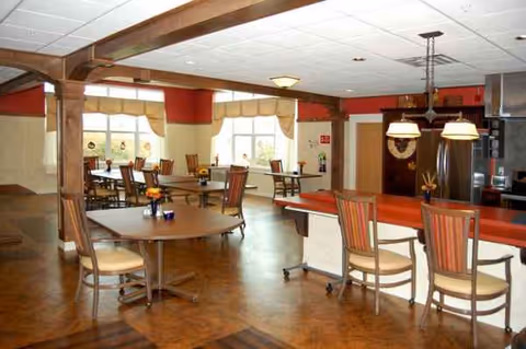 Interior dining area with round tables, chairs, and a red counter in a senior living facility.