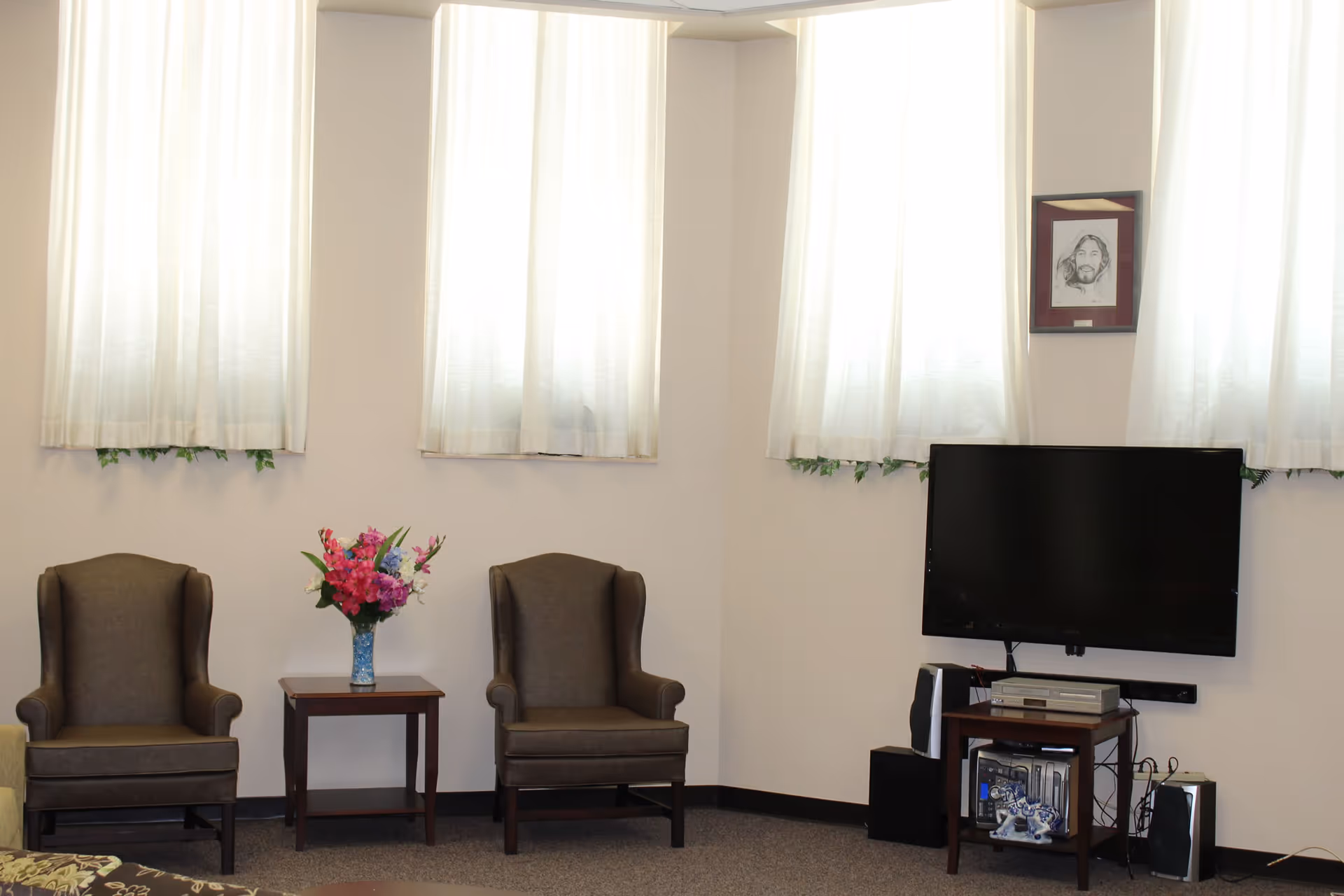 A cozy living room area with two brown armchairs separated by a small wooden table holding a vase of colorful flowers. On the right side, a flat-screen TV is mounted on the wall above a small wooden stand with electronic devices and speakers. The room has three tall windows with white curtains allowing natural light to fill the space. A framed sketch is hung on the wall above the TV.