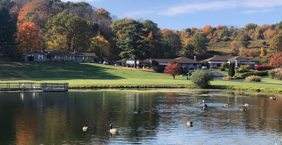 A scenic view of Valley Vista Assisted Living facility with a pond in the foreground where several ducks are swimming. The background shows a grassy area with trees displaying autumn colors and multiple single-story buildings.