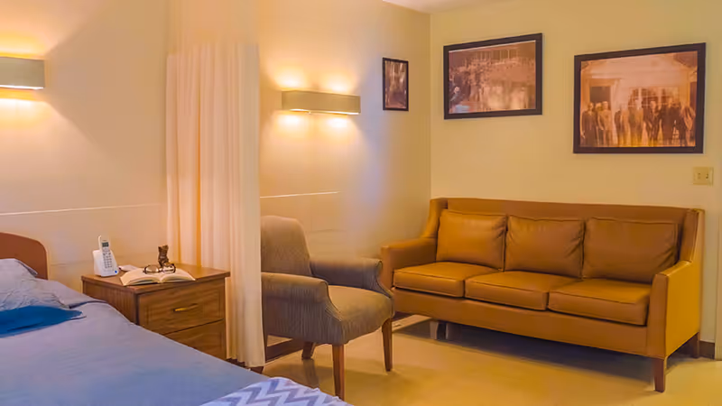 A cozy room with a bed on the left side, a wooden nightstand with a telephone and an open book, a gray upholstered armchair, and a brown leather sofa against a wall decorated with three framed pictures. Soft wall-mounted lights illuminate the space.